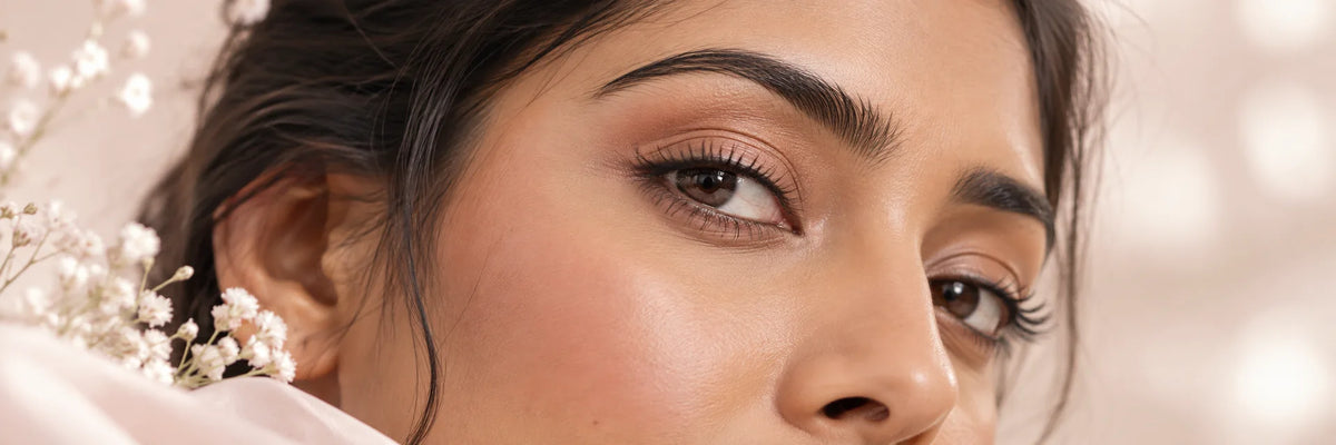 An editorial close-up of one adult South Asian woman’s relaxed eye and upper cheek with natural healthy skin texture, gentle neutral makeup, soft ivory and blush studio lighting, no balm or applicator.