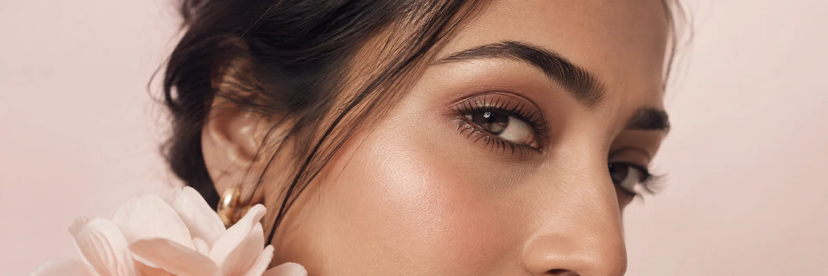 An editorial close-up of one adult South Asian woman’s eye and temple, understated softly defined lash line and natural skin texture, pale blush studio background