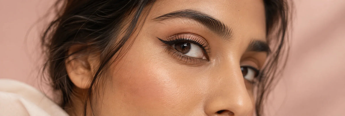 An editorial close-up of one adult South Asian woman’s eye with a refined thin black winged eyeliner and realistic natural skin, blush studio background, no applicator.
