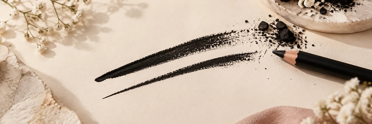 Two precise rich black kajal strokes on warm ivory paper alongside subtle charcoal pigment texture, premium graphic editorial beauty still life.