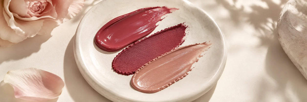 A richly glossy rose lip-colour texture beside matte berry and satin nude swatches on ivory ceramic, beautiful editorial colour still life.