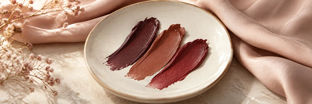 Deep plum, warm cinnamon and wine lipstick swatches on ivory ceramic with soft blush silk, sophisticated warm colour composition.