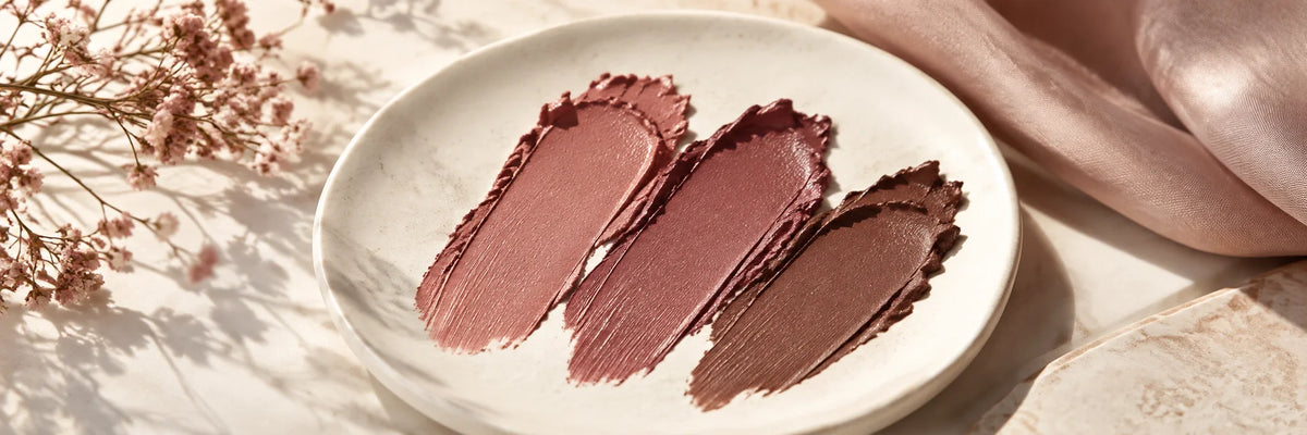 Muted rosewood, rich mauve and warm cocoa lipstick textures on ivory ceramic, refined neutral beauty colour palette.