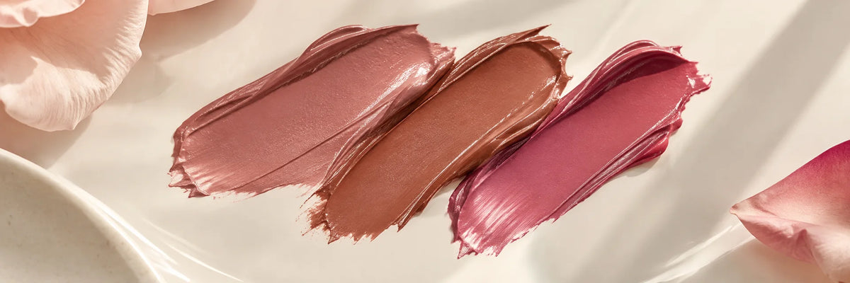 Smooth luminous satin lipstick strokes in mauve, cinnamon and soft fuchsia on ivory ceramic, subtle satin reflections.