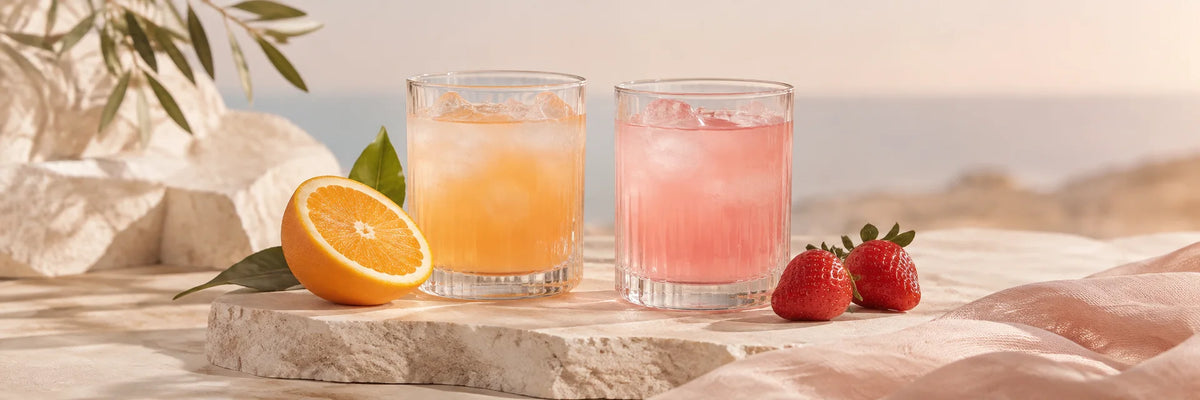 Two clear water glasses with pale orange and pale pink beverages, a sliced orange and two strawberries beside them on ivory stone, fresh understated blush daylight, no pills or supplement bottles.